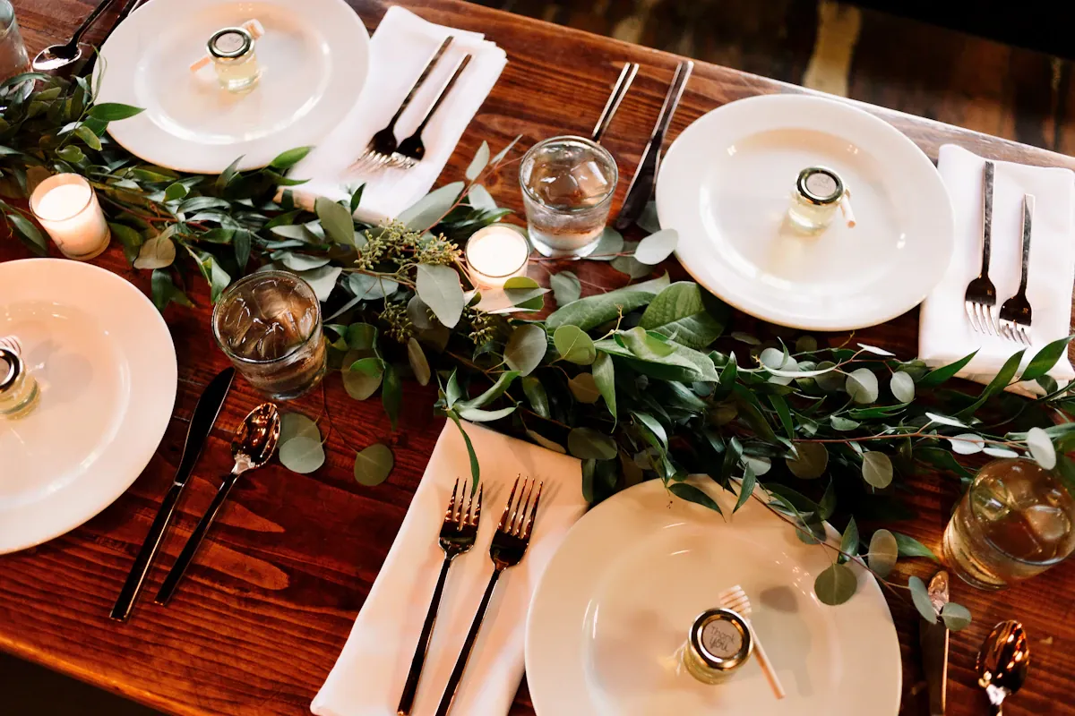 White plates, glassware, cutlery and greenery arranged on a wooden dining table