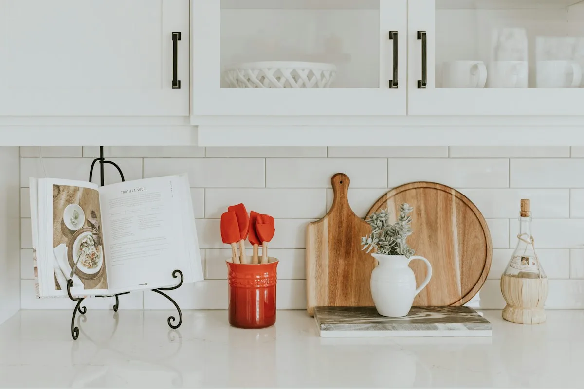 White kitchen counter with an open cookbook, wooden boards and a utensil pot