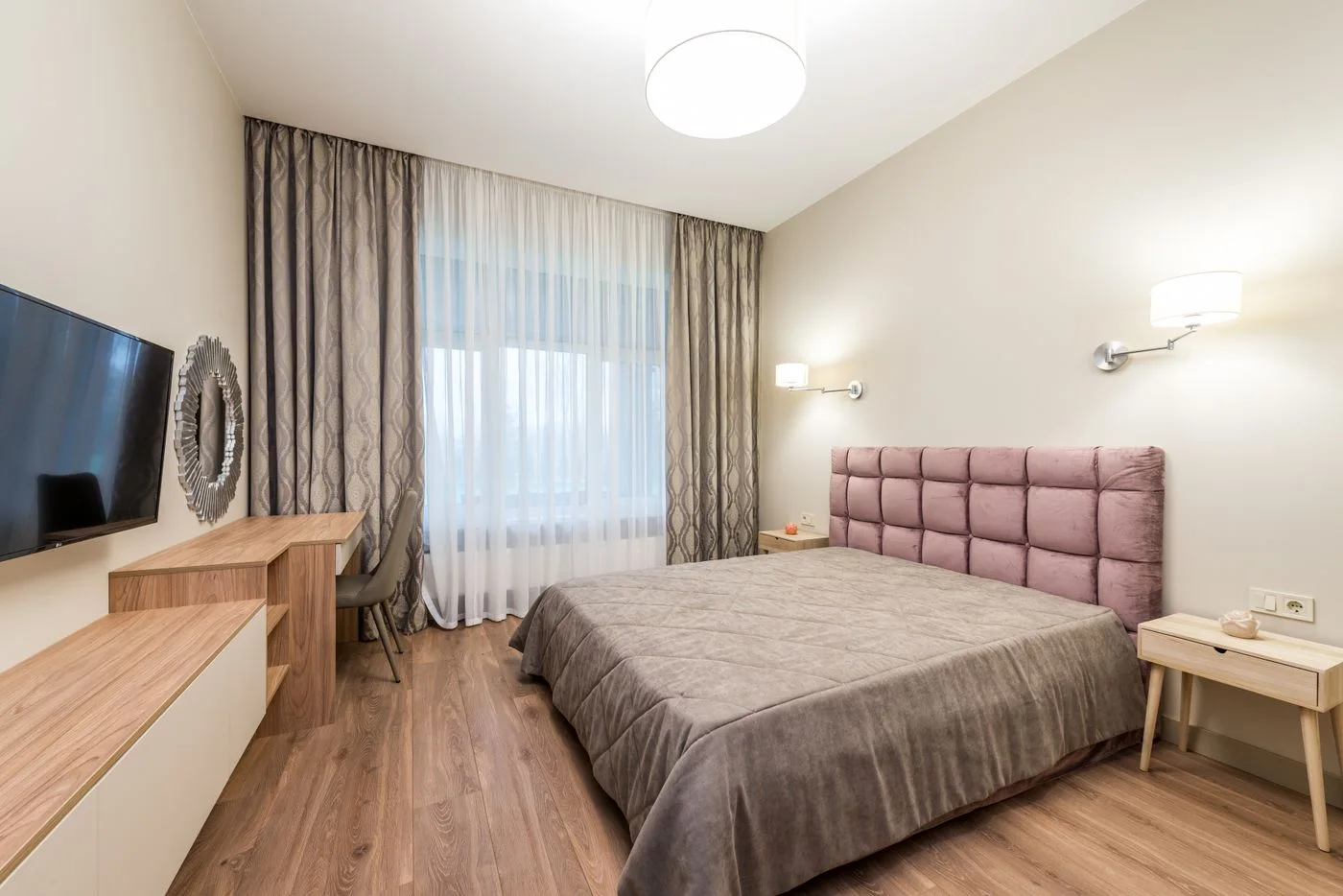 A bedroom with upholstered headboard, window curtains and a wall screen