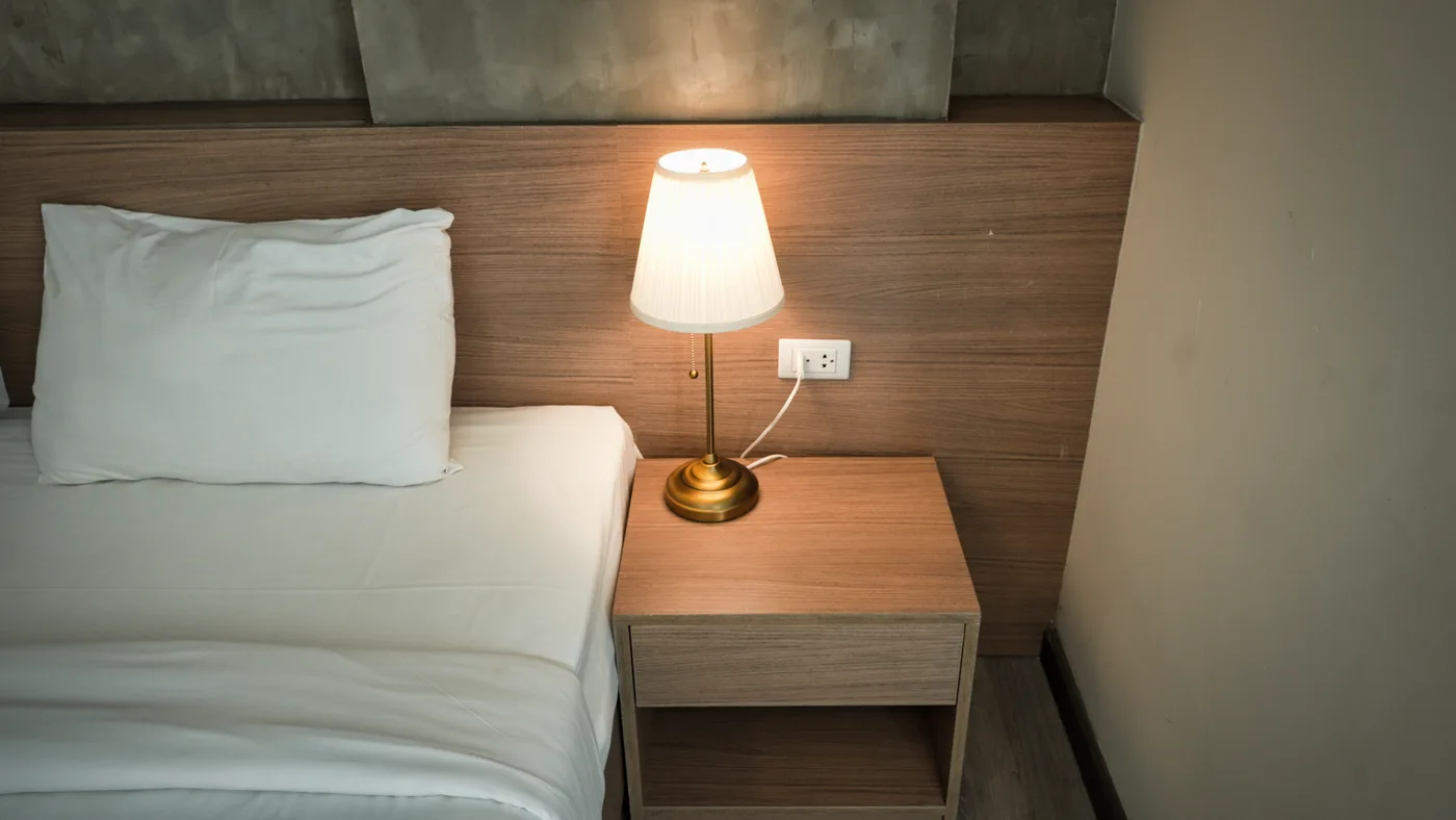 A bedside lamp on a wooden table beside pale bedding