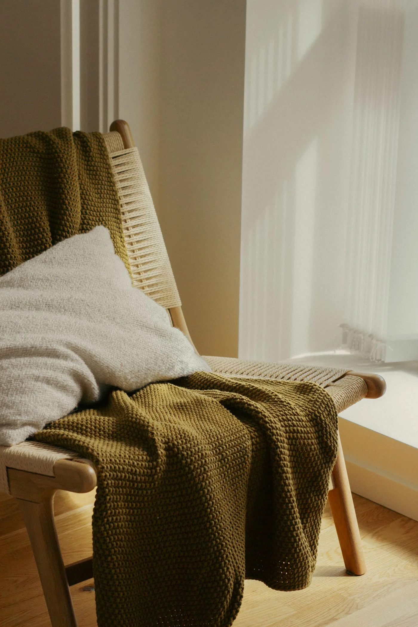 An upholstered chair with a cushion and textured throw in window light